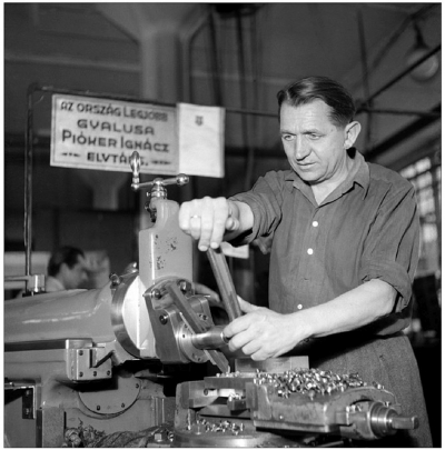 Ein Beispiel für die visuelle Inszenierung von Ignác Pióker als Held der sozialistischen Arbeit durch die Agentur MTI. Pióker wurde vornehmlich bei der Arbeit oder während der Ausbildung des Hobler-Nachwuchses dargestellt. Zudem entstanden Home-Storys mit seiner Ehefrau und den beiden Söhnen. An Klarheit ist diese Komposition kaum zu übertreffen: In der Mitte des Bildes befindet sich die rechte Hand des Hoblers, also der Teil des Körpers, dem die besonders präzise – und aus diesem Grund mehrfach ausgezeichnete – Arbeit zugeschrieben wurde. Der Fokus des Betrachters richtet sich auf die Maschine sowie den Mann dahinter. Etwas weiter links im Bild ist ein Schild zu erkennen mit der Aufschrift: »Der beste Hobler des Landes, Genosse Ignácz Pióker«. (MTI-Fotoarchiv/Magyar Fotó, Nr.: FMAFI19509156002, Fotograf: Ferenc Bartal)