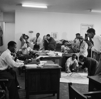 Die Redaktion bei der Arbeit, 1954: »The overcrowded Drum office in Johannesburg housed most of its journalists and photographers. Jurgen Schadeberg took the picture while Anthony Sampson directed it, showing (from left to right) Henry Nxumalo, Casey Motsitsi, Ezekiel Mphalele, Can Themba, Jerry Ntsipe, Arthur Maimane (wearing hat, drooping cigarette), Kenneth Mtetwa (on floor), Victor Xashimba, Dan Chocho (with hat), Benson Dyanti (with stick) and Robert Gosani (right with camera). Todd Matshikiza was away.« (Zitat: <http://www.jurgenschadeberg.com/galleries/TheBlack_WhiteFiftiesinSouthAfrica/BW50s_34.htm>; Foto: Jürgen Schadeberg)