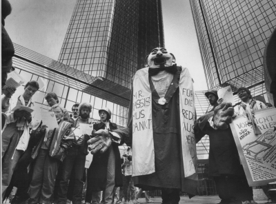 Die Figur des »Mr. Apartheid« bzw. »Mr. Rassismus« wurde von westdeutschen Anti-Apartheid-Aktivisten in den 1980er-Jahren entworfen und begleitete deren Kampagne gegen Banken, die Kredite nach Südafrika vergaben – hier bei einer Protestaktion im Frankfurter Bankenviertel während des Deutschen Evangelischen Kirchentags im Juni 1987. In dieser Darstellung wurde das Apartheid-System zu einer monströsen Gestalt mit riesigem Mund und übergroßen Händen stilisiert, die durch ausländisches Kapital am Leben erhalten wird. Der Text auf der Schärpe lautete vermutlich »Mr. Rassismus dankt für die Kredite aus der BRD«. Ähnlichkeiten mit dem südafrikanischen Präsidenten Pieter Willem Botha waren wohl beabsichtigt. Nach dem Ende der Apartheid wurde die Puppe nach Südafrika verschenkt und begrüßt heute Besucher von Robben Island im Eingangsbereich des Nelson Mandela Gateways in Kapstadt. (Institut für Stadtgeschichte, Frankfurt a.M., Signatur: ISG_S7FR_6096, Foto: Luigi Ungarisch)