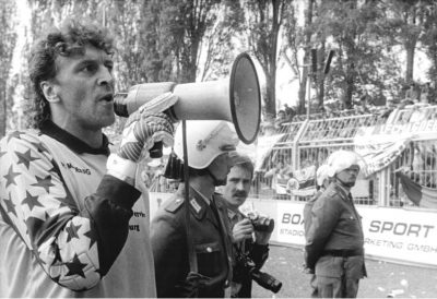 26. Mai 1990, Fußball-Oberliga der DDR: Krawalle auf der Zuschauertribüne der Gäste aus Magdeburg überschatteten das Match im Dr.-Kurt-Fischer-Stadion in Karl-Marx-Stadt (ab Juni 1990 wieder Chemnitz). Nachdem Feuerwerkskörper von den Rängen geflogen waren und das Spiel daraufhin unterbrochen werden musste, rief Magdeburgs Torwart Dirk Heyne die Fans per Megaphon zur Ruhe auf.(Bundesarchiv, Bild 183-1990-0526-010, Foto: Jan Peter Kasper) 26. Mai 1990, Fußball-Oberliga der DDR: Krawalle auf der Zuschauertribüne der Gäste aus Magdeburg überschatteten das Match im Dr.-Kurt-Fischer-Stadion in Karl-Marx-Stadt (ab Juni 1990 wieder Chemnitz). Nachdem Feuerwerkskörper von den Rängen geflogen waren und das Spiel daraufhin unterbrochen werden musste, rief Magdeburgs Torwart Dirk Heyne die Fans per Megaphon zur Ruhe auf. (Bundesarchiv, Bild 183-1990-0526-010, Foto: Jan Peter Kasper)